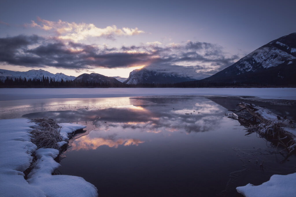 Image of a winter sunrise in Banff national park. Frozen lake with reflection of a mountain in the background as the sun peaks around it.