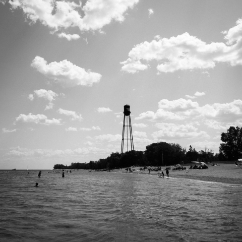 Black and white photograph of Winnipeg beach. People play on the beach and in the water with a water tower in the background