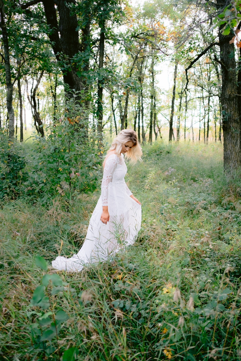 A bride in a white dress in a forest looking down at grass.