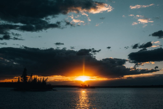 Photo of a cloudy sunset over the lake with the sun backlighting an island in the distance.